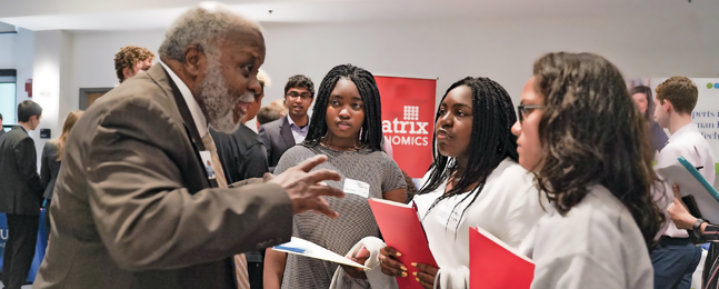 A man with gray hair and beard talks animatedly to three young women.